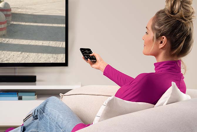 Person auf Couch steuert mit Handy den Fernseher.